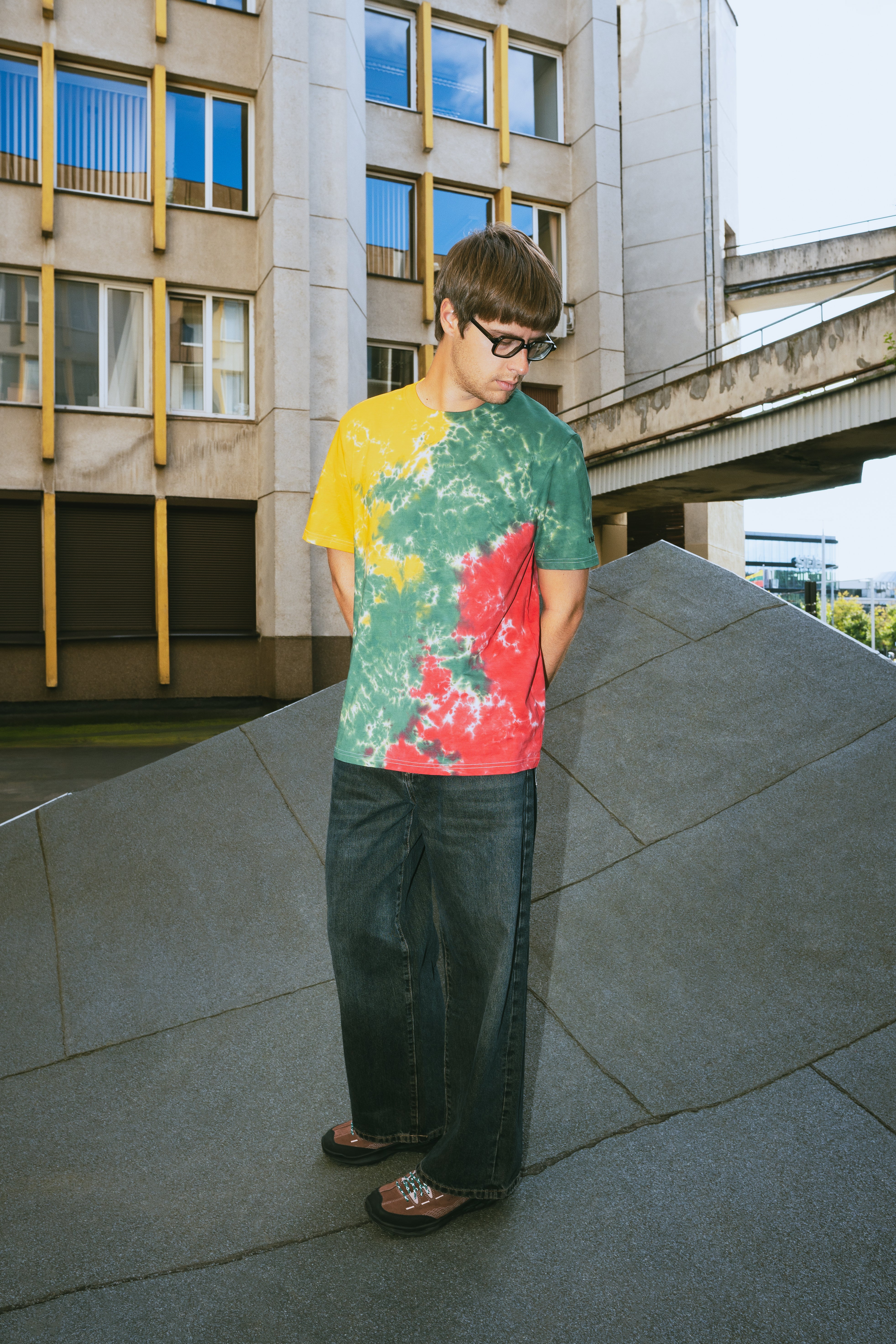 A man wearing a colorful tie-dye t-shirt with yellow, green, and red sections, paired with dark wide-leg jeans and sandals, standing outdoors on a concrete structure in front of a modern building.