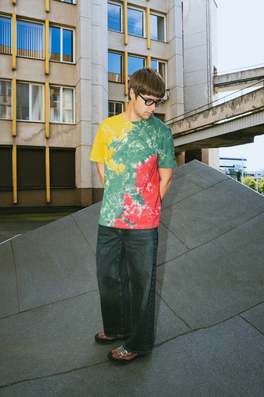 A man wearing a colorful tie-dye t-shirt with yellow, green, and red sections, paired with dark wide-leg jeans and sandals, standing outdoors on a concrete structure in front of a modern building.