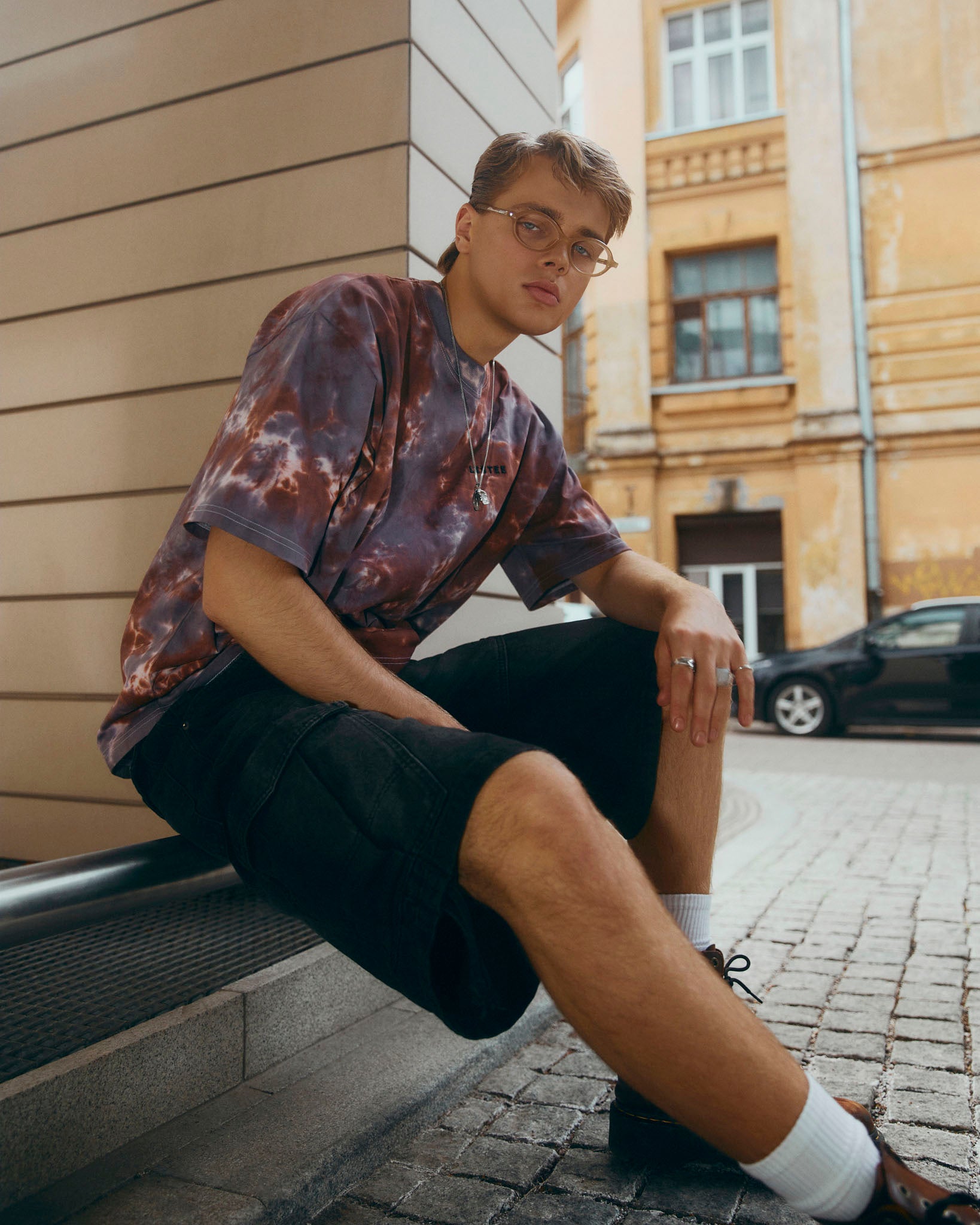A young man wearing a brown tie-dye T-shirt, black cargo shorts, white socks, and brown leather shoes sits on a stone ledge in an outdoor urban setting.