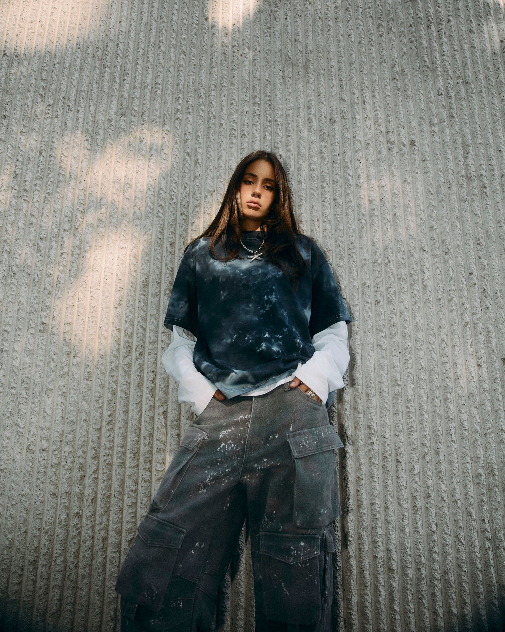 A woman stands against a textured concrete wall wearing a dark grey and black tie-dye t-shirt layered over a white long-sleeve shirt, paired with dark gray cargo pants.