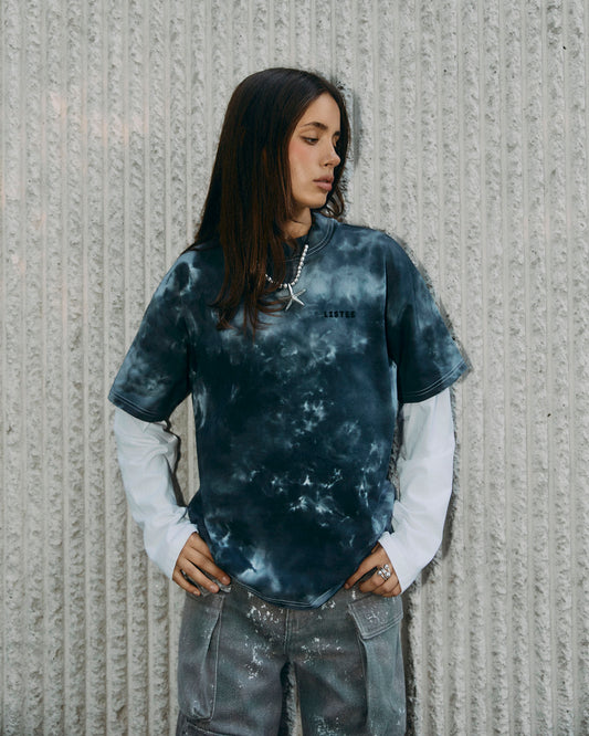 A woman wearing a dark grey and black tie-dye t-shirt with a white long-sleeve shirt underneath, standing against a textured gray concrete wall.