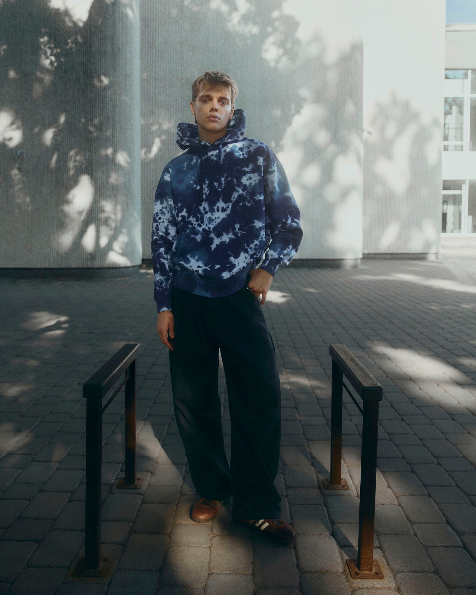 A man wearing a navy blue and light blue tie-dye hoodie, dark wide-leg trousers, and brown leather shoes stands outdoors on a paved walkway between two black metal bike racks.