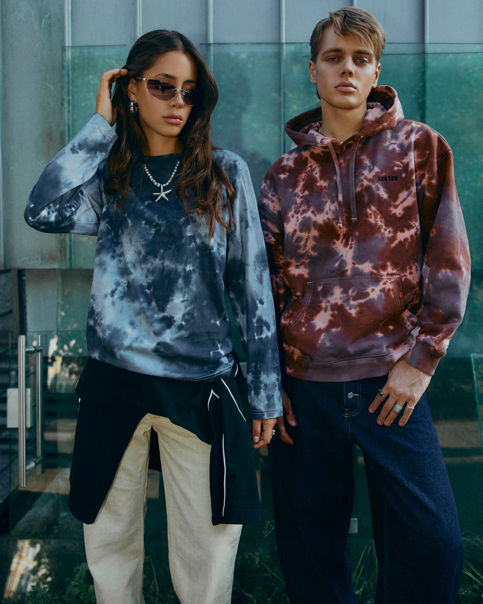 Two models wearing tie-dye sweatshirts in a modern outdoor setting with glass walls. The model on the left wears a slate black and grey tie-dye long-sleeve T-shirt with cream pants. The model on the right wears a brown tie-dye hoodie.