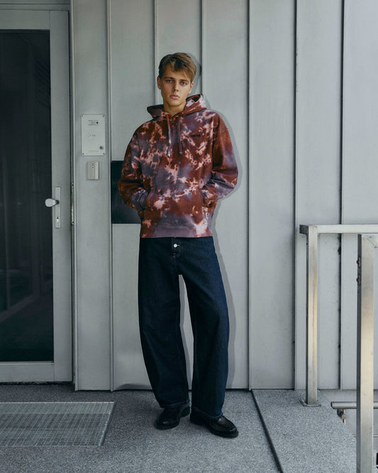 A model stands on a concrete porch wearing a brown tie-dye hoodie, dark blue jeans, and black loafers, set against a gray paneled wall.