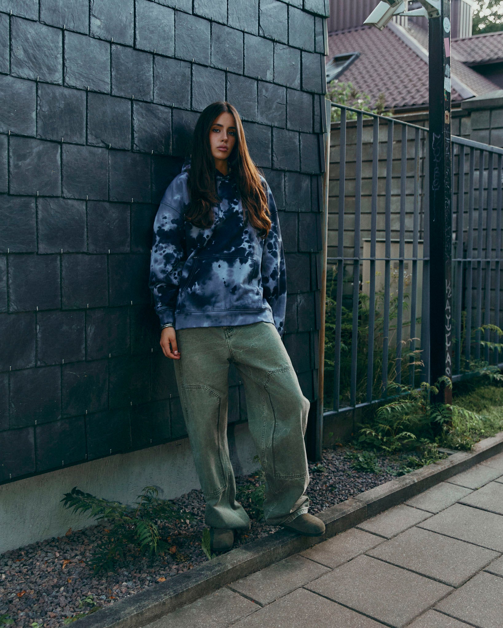 A woman stands against a dark gray brick wall in an outdoor urban setting, wearing a grey and black tie-dye hoodie, loose olive green pants, and dark boots.