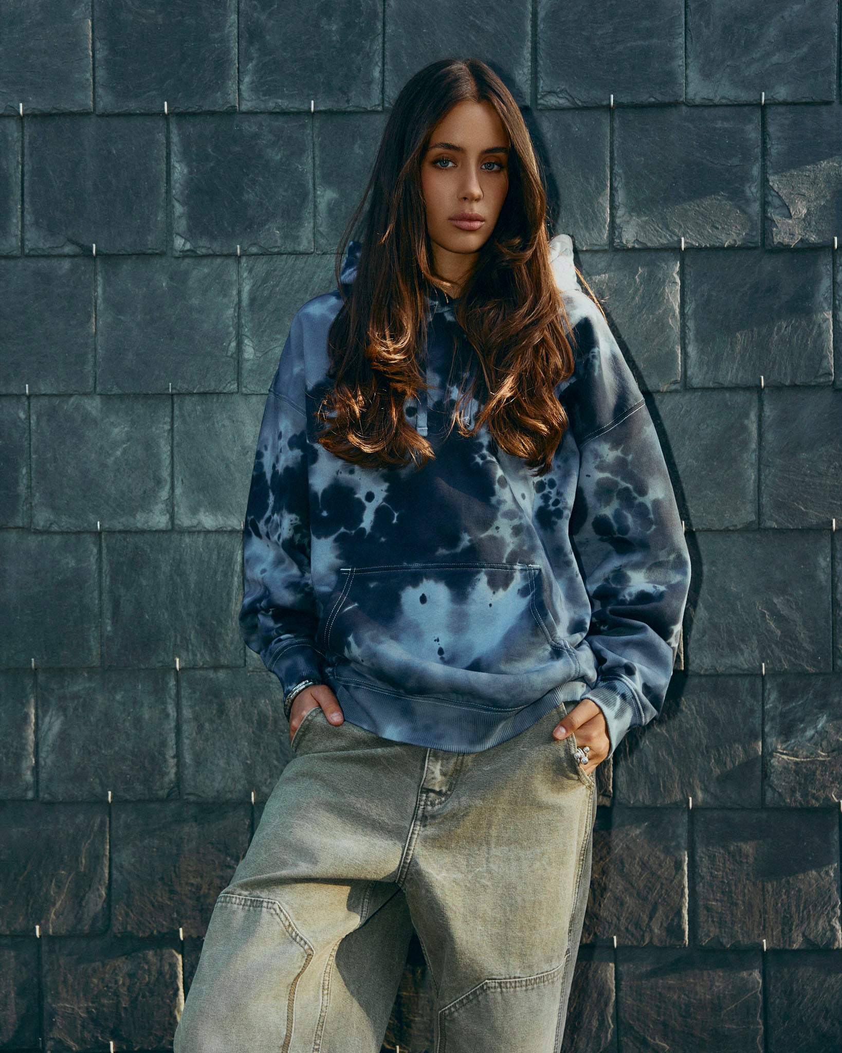 A black and grey tie-dye pullover hoodie with a small black text logo on the left chest, displayed against a solid light gray background.